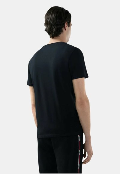 Uomo con capelli scuri che indossa una maglietta nera a maniche corte e pantaloni neri con dettaglio di logo a strisce bianche e rosse, visto di spalle su sfondo semplice.