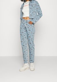 Chaqueta de mezclilla azul claro con estampado floral, combinada con unos pantalones de flores de cintura alta a juego. Top corto blanco y zapatillas gruesas blancas.