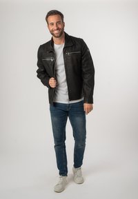 Giacca di pelle nera con tasche con zip, indossata sopra una maglietta grigia chiara. Abbinata a jeans blu e sneakers bianche. In piedi su uno sfondo chiaro.