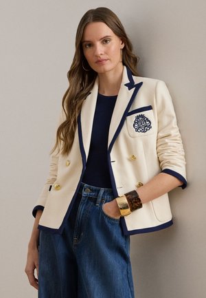 Blazer crème avec bordure marine, boutons dorés croisés et poche poitrine brodée. Porté sur un top marine et associé à un jean.