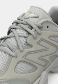 Gros plan d'une basket New Balance 9060 gris clair montrant un tissu en mesh, une languette rembourrée, un grand logo N et des lacets noués.
