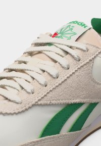 Κοντινό πλάνο μπεζ και λευκού αθλητικού παπουτσιού Reebok με λευκά κορδόνια, πράσινες ρίγες στο πλάι και πράσινο λογότυπο στη γλώσσα.