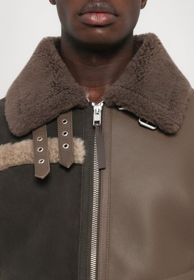 Braune Lederjacke mit einem flauschigen Schaffellkragen, Reißverschluss und kontrastierenden braunen Patches. Mit Metallbeschlägen und Nähten verziert.
