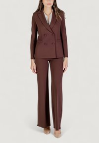 Hnědý dvouřadý blazer s dvěma předními kapsami, spárovaný s odpovídajícími rozšířenými kalhotami. Texturovaná tkanina, střih na míru a světlá košile pod tím.