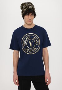 Jūras zila kokvilnas T-krekls ar lielu zelta Versace Jeans Couture logotipu apļa dizainā. Komplektā ar melnu un zelta cepuri.