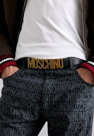 Černý opasek s výrazným zlatým logem "MOSCHINO", spárovaný s tmavými vzorovanými kalhotami s opakujícím se textem. Doplněný o bílou košili a hnědou bundu.