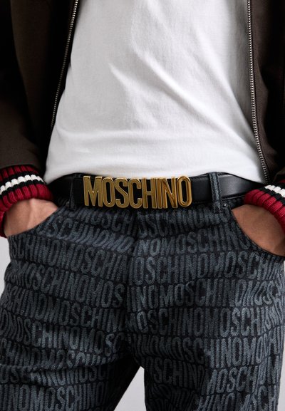 Mustaräbelt, millel on silmapaistev kuldne "MOSCHINO" logo, koos tumedate mustriliste pükstega, millel on korduv tekst. Kands mütsi valget särki ja pruuni jakki.