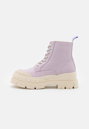 Lavendelfarbener Canvas-Stiefelette mit dicker beiger Profilsohle, Schnürung vorne und Zuglasche hinten, Seitenansicht vor weißem Hintergrund.