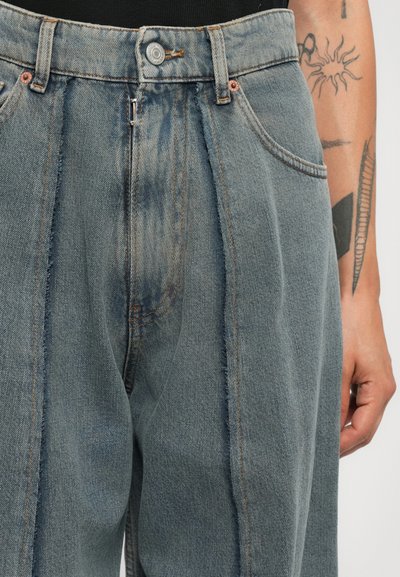 Vedere frontală a blugilor gri din denim cu talie înaltă, cu plisuri, închidere cu buton și un braț tatuat al unei persoane odihnindu-se lângă corp.