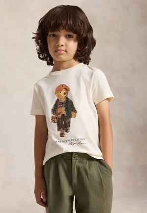 Barn med mellem-langt krøllet hår iført en hvid t-shirt med en bjørnegrafik og grønne bukser, stående mod en neutral baggrund.