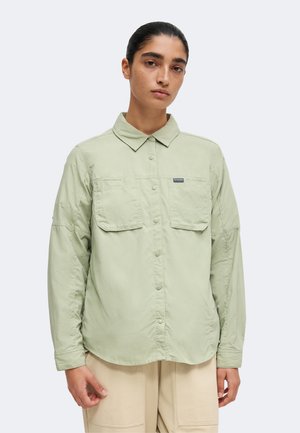 Persona con cabello oscuro y camisa verde claro abotonada con dos bolsillos en el pecho, de pie frente a un fondo claro y sencillo.