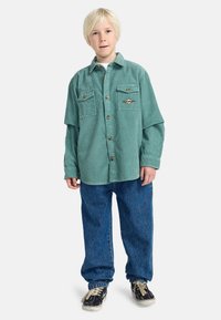 Groene corduroy shirt met twee borstzakken, houten knopen en een decoratieve badge, gecombineerd met losse blauwe denim broek en casual sneakers.
