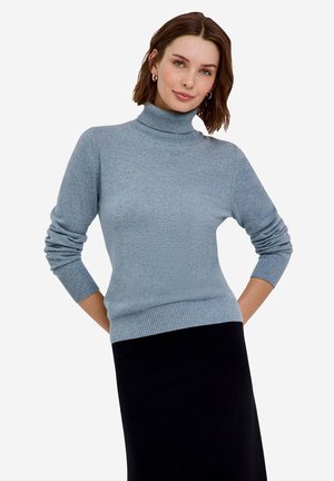 Femme aux cheveux courts et bruns, portant un pull à col roulé bleu clair et une jupe noire, debout avec les mains dans le dos sur fond blanc.