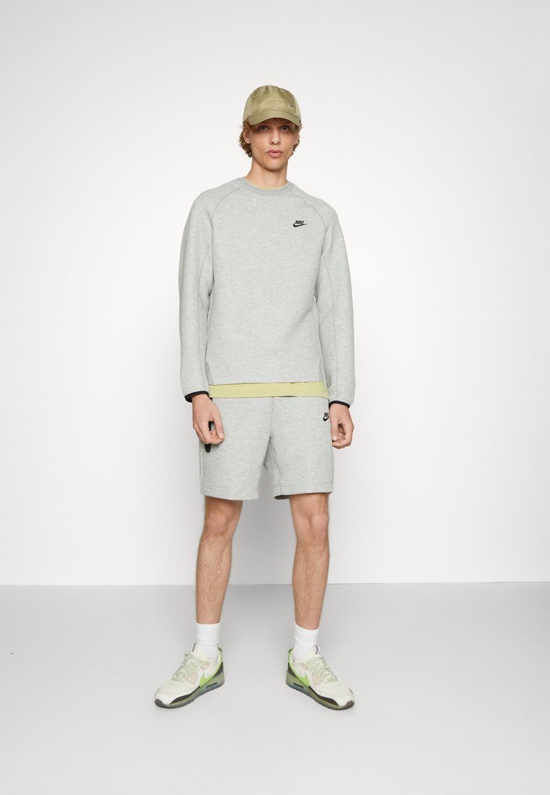 Sweat à capuche gris avec logo noir, shorts assortis et bandeau vert olive clair. Associé à une casquette beige et des sneakers avec des accents verts.