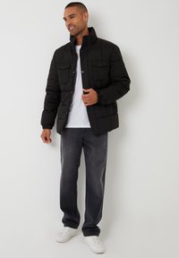 Schwarze Daunenjacke mit zwei Brusttaschen, Druckknöpfen und elastischen Bündchen, kombiniert mit grauen Jeans und weißen Turnschuhen. Darunter ein schlichtes weißes T-Shirt.