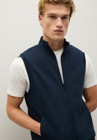 Gilet senza maniche blu navy realizzato in materiale morbido, con zip frontale, colletto alto e tasche laterali, indossato sopra una maglietta bianca.