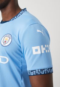 Vaaleansininen urheilupaita, jossa on pyöreä kaulus ja teksturoitu pinta. Rinnassa on Manchester Cityn logo ja olkapäällä Puman logo.