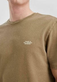 Homme portant un t-shirt marron avec un texte blanc sur la poitrine indiquant "PRENEZ UNE PAUSE BIEN MÉRITÉE" sur fond uni.
