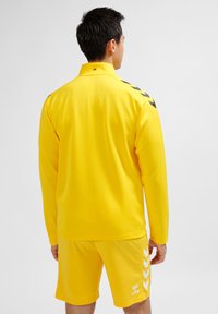 Gul idrottsjacka och shorts i mjukt, lättviktigt material. Har svarta chevrondetaljer på axlarna och shorts.