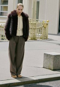 Chaqueta de pelo sintético burdeos, blusa crema con cinturón, pantalones anchos de cuadros marrones, mocasines en tonos neutros. Entorno urbano con un bordillo de piedra.