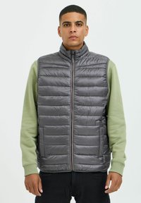 Chaleco acolchado gris con cuello alto, que presenta paneles acolchados horizontales y un cierre frontal. Llevado sobre una camiseta de manga larga verde.
