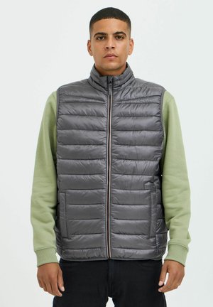 Gilet rembourré gris avec un col montant, présentant des panneaux matelassés horizontaux et une fermeture éclair sur le devant. Porté par-dessus un haut à manches longues vert.