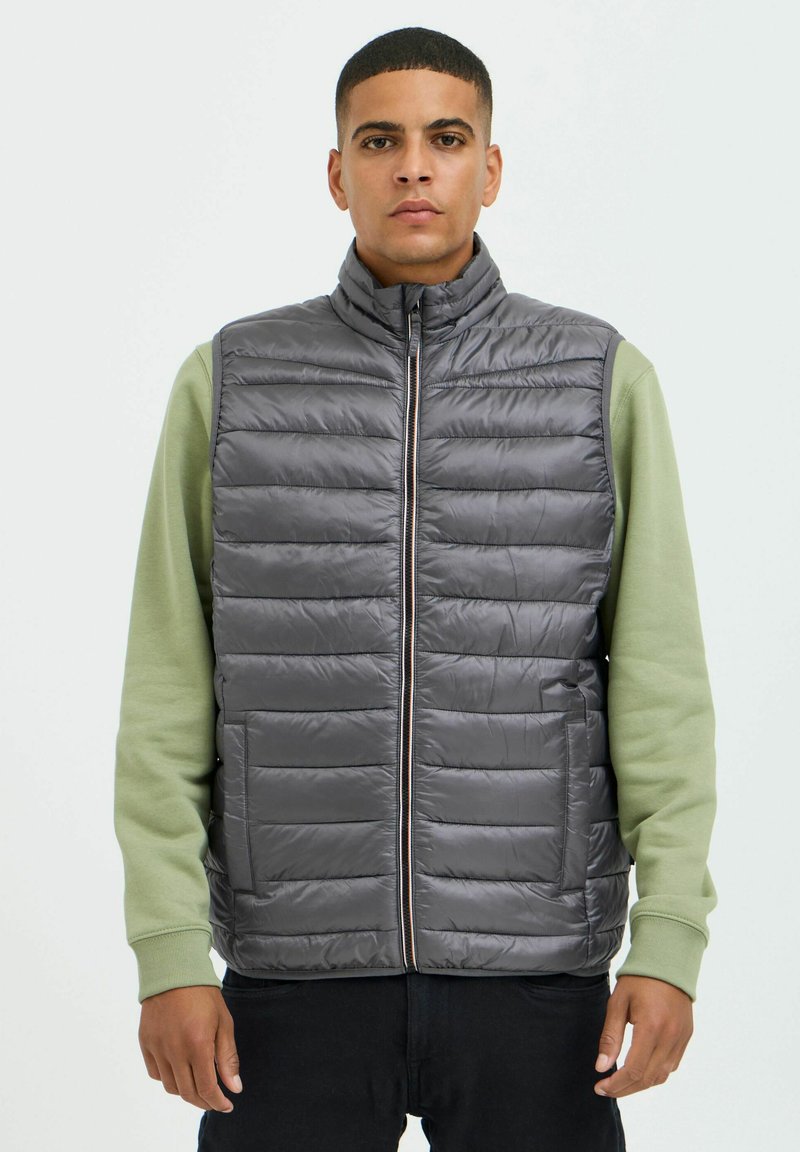 Chaleco acolchado gris con cuello alto, que presenta paneles acolchados horizontales y un cierre frontal. Llevado sobre una camiseta de manga larga verde.