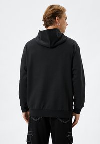 Svart huvtröja med en avslappnad passform, som har en mjuk textur och ribbade ärmslut och nederkant. Bakifrån visas en sömlös huva.