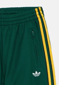 Grønne joggebukser i mesh med en teksturert overflate, med gule og svarte sidestriper, elastisk midjebånd og glidelåslommer. Hvitt logodetalj.