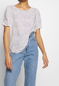 Mujer con una blusa blanca de manga corta con puntos negros y jeans de tiro alto de color azul claro, contra un fondo liso.