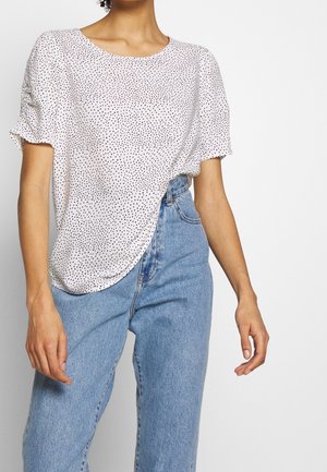 Kvinna som bär en vit kortärmad blus med svarta prickar och högmidjade ljusblå jeans mot en neutral bakgrund.
