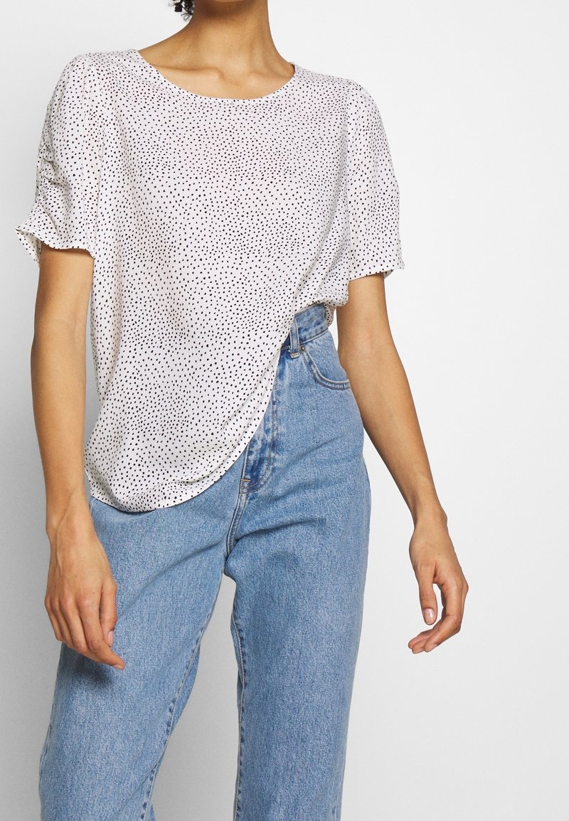Mujer con una blusa blanca de manga corta con puntos negros y jeans de tiro alto de color azul claro, contra un fondo liso.