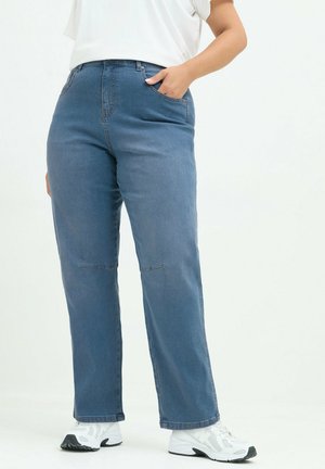 Personne portant un jean bleu droit taille haute, des baskets blanches et un haut blanc, avec une main dans la poche, sur un fond uni.