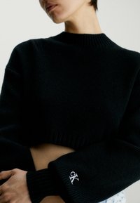 Schwarzer gerippter, kurz geschnittener Strickpullover mit hohem Kragen und weiten Ärmeln. Verfügt über ein kleines weißes "CK"-Logo am linken Bündchen.