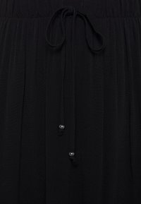 Sort kjole med plisseret tekstur, der har snøre i taljen og metalliske perledekorationer på snørene. Blødt, letvægtsstof.