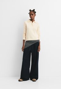 Femeie cu coafura bantu knot, purtând un pulover crem, o curea gri, pantaloni negri largi și pantofi bej, stând în fața unui perete alb.