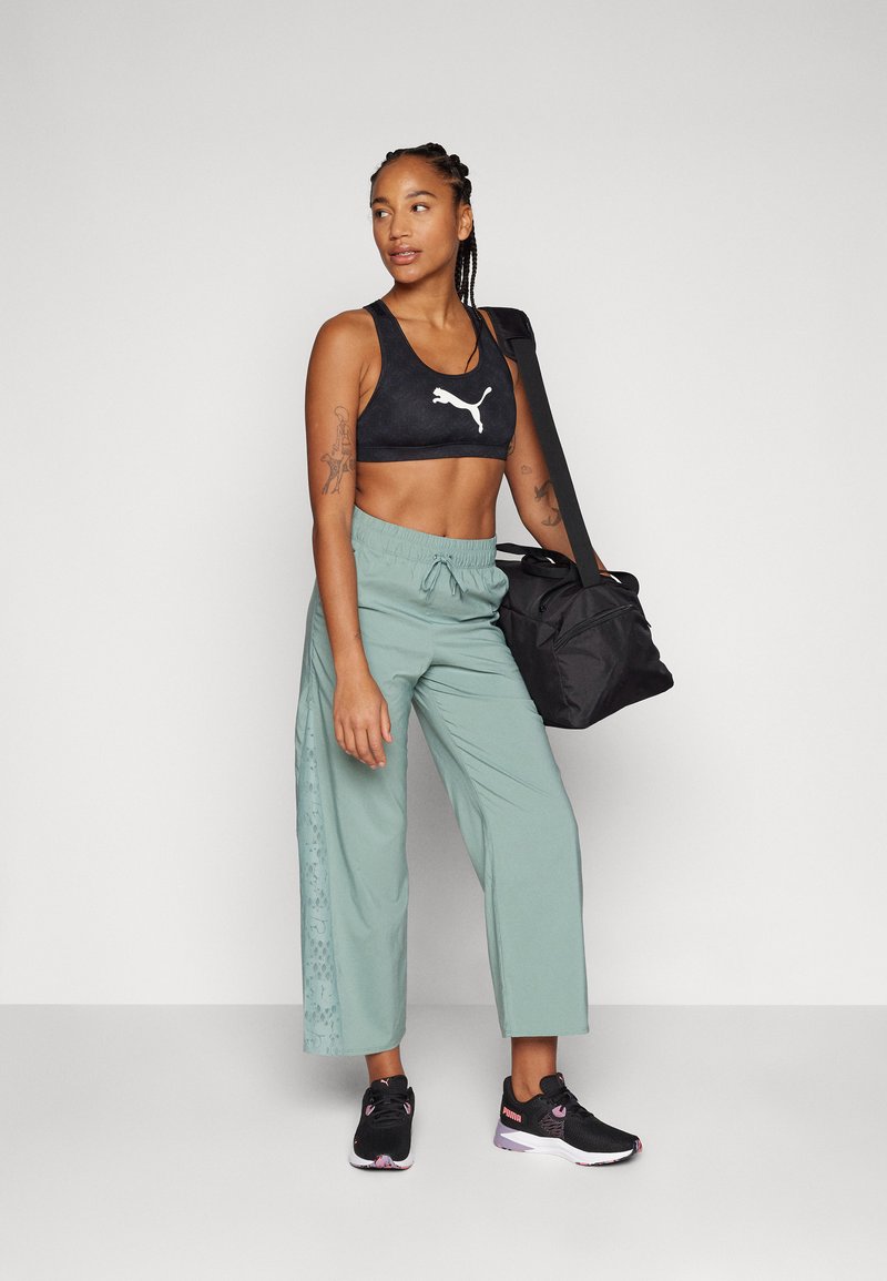 Reggiseno sportivo nero con logo Puma, pantaloni verde chiaro con dettagli in pizzo laterali, sneakers nere, in possesso di una borsa a tracolla nera.