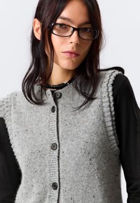 Jeune femme aux cheveux bruns et portant des lunettes, vêtue d'un gilet en tricot gris à boutons sur un t-shirt noir à manches longues, devant un fond uni.