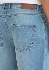 Jeans de mezclilla azul claro con costuras visibles, un parche de cuero en la parte trasera con el nombre de la marca y un diseño estándar de bolsillo trasero.
