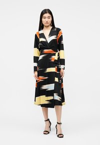 Γυναίκα που φοράει μαύρο φόρεμα τύπου wrap με μακριά μανίκια και αφηρημένα μοτίβα με πινελιές σε κίτρινο, πορτοκαλί, λευκό και ανοιχτό γαλάζιο, συνδυασμένο με μαύρα ψηλοτάκουνα.