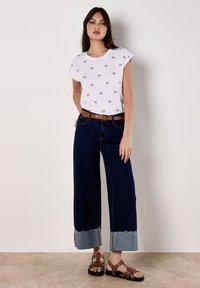 La modelo se presenta con confianza en una camiseta blanca con pequeños estampados de lazo, combinada con unos vaqueros de pierna ancha y sandalias marrones, todo ello frente a un fondo neutro.