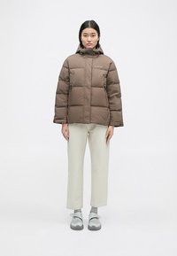 Smeđi puffer jakna s patentnim zatvaračem, kapuljačom i nabranim kvadratima. Nosena preko svijetlih hlača i sivih cipela. Minimalistički dizajn.