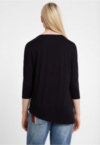 Kvinna med långt blont hår klädd i en lös svart topp med trekvartsärmar och blå jeans med en rutig lapp, som står mot en enkel vit bakgrund.