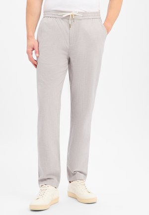 Homme portant un pantalon gris clair à cordon et des baskets blanc cassé, debout les mains dans les poches devant un fond blanc.