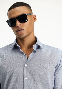 Camicia a fantasia blu con colletto button-down, caratterizzata da piccoli disegni geometrici; occhiali da sole quadrati neri con montatura lucida.