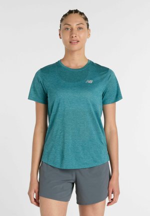 Femme aux cheveux tressés portant un t-shirt de sport à manches courtes bleu sarcelle et un short gris, debout devant un fond blanc uni.