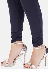 Marineblaue Leggings verfügen über gerafften Stoff am Knöchel und sind kombiniert mit glänzenden silbernen High-Heelsandalen mit Riemen und offenem Zehendesign.