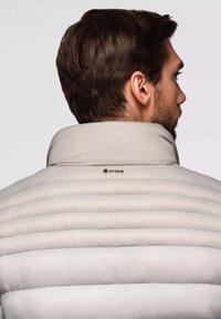 Hellgraue Pufferjacke mit vertikaler Steppung, hohem Kragen und einem Logopan über dem Nackenbereich. Geschmeidige Textur, isoliertes Design.