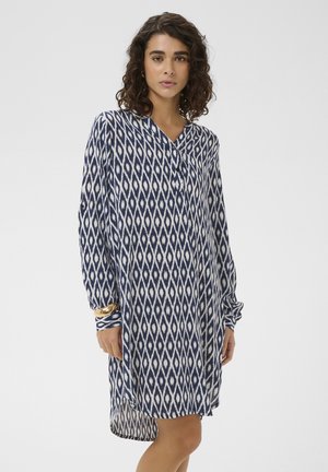 Woman wearing a long-sleeve navy and white diamond patterned dress with a V-neck and gold bracelet on left wrist, standing against a plain background.