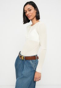 Μπλούζα με μακριά μανίκια σε εφαρμοστή γραμμή από εκρού ριμπ ύφασμα, με λαιμόκοψη henley. Φορεμένη με μπλε τζιν παντελόνια ψηλόμεσα και καφέ ζώνη.